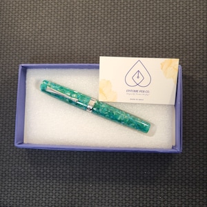 May include: A teal and white marbled fountain pen with silver accents rests in a blue box with white padding. A business card with the logo of "Epitome Pen Co." is also in the box. The pen is a writing instrument.