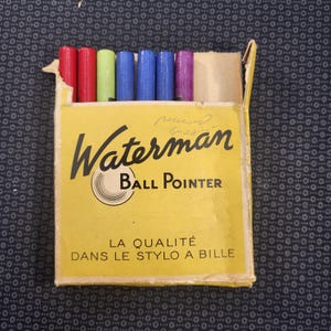 May include: A vintage yellow Waterman Ball Pointer pen box with a partial view of the colorful pens inside. The box has the text "Waterman Ball Pointer" and "LA QUALITÉ DANS LE STYLO A BILLE". The pens are red, green, blue, and purple.