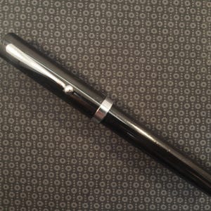 May include: A black fountain pen with silver accents. The pen has a glossy finish and a silver band near the center. A small silver detail is visible near the top of the pen.