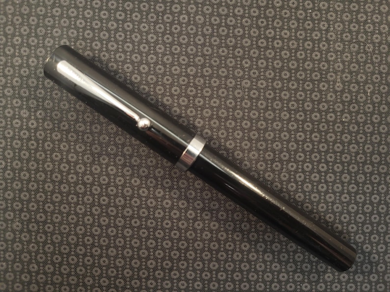 May include: A black fountain pen with silver accents. The pen has a glossy finish and a silver band near the center. A small silver detail is visible near the top of the pen.