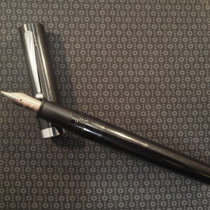 May include: A black fountain pen with a silver cap and nib. The pen is lying on a patterned surface. The pen's nib is silver and gold-toned. The pen is a writing instrument.