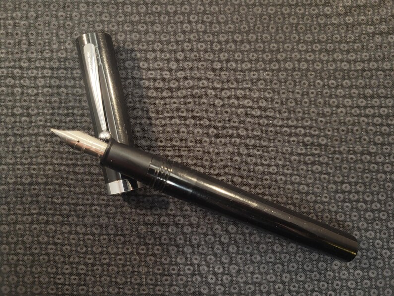 May include: A black fountain pen with a silver cap and nib. The pen is lying on a patterned surface. The pen's nib is silver and gold-toned. The pen is a writing instrument.