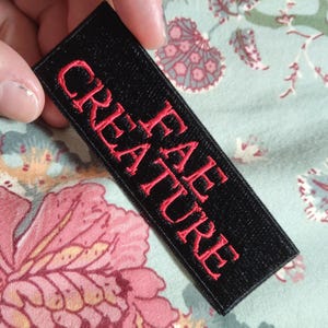 Puede incluir: Un parche rectangular negro con las palabras "CREATURE FAE" bordadas en rojo. El parche se sostiene sobre un fondo con estampado floral.