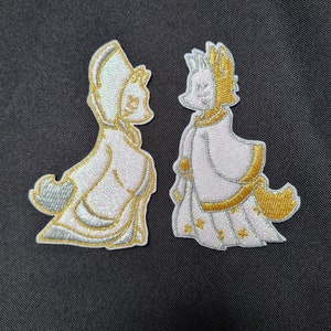 May include: Two embroidered iron-on patches featuring a white fox wearing a pink and gold robe. The fox on the left is facing right and the fox on the right is facing left.