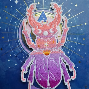 May include: Embroidered patch featuring a stylized beetle in shades of pink, purple, and orange, with white outlining. The patch is set against a dark blue background with celestial-themed gold accents, including stars and a crescent moon.