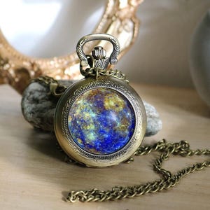 May include: Vintage brass pocket watch with a chain. The watch face displays a blue, yellow, and green pattern. The watch is on a wooden surface, with a gold frame and a small plant in the background.