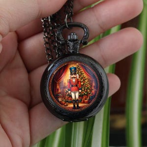 Könnte beinhalten: Eine Taschenuhr aus schwarzem Metall mit einem runden Gehäuse und einer passenden Kette. Das Zifferblatt zeigt eine farbenfrohe Illustration eines Nussknacker-Soldaten, der vor einem geschmückten Weihnachtsbaum mit Geschenken steht.