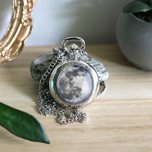 Peut inclure: Un montre de poche argentée avec une chaîne, représentant une image détaillée de la lune. La montre a un design orné autour du cadran et une boucle en haut pour la chaîne. La chaîne est posée sur une pierre grise.
