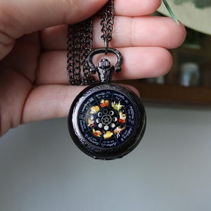 Puede incluir: Colgante de reloj de bolsillo negro con una cadena a juego. La esfera del reloj presenta un diseño del zodíaco con ilustraciones coloridas y texto. El reloj se sostiene en una mano.