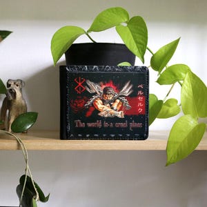 May include: Black leather wallet with a graphic of a character from the anime Berserk. The text "The world is a cruel place" and Japanese text are printed on the wallet.