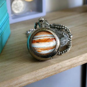 Puede incluir: Un reloj de bolsillo plateado con cadena, que presenta una imagen detallada de Júpiter. La esfera del reloj muestra las bandas anaranjadas, blancas y azules del planeta. El reloj está sobre una superficie de madera.