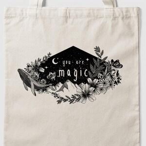 Könnte beinhalten: Eine beigefarbene Stofftasche mit einem schwarzen Grafikdruck, der einen Wal, Berge, Blumen und einen Schmetterling zeigt, die eine nächtliche Himmelsszene mit Mondsichel und Sternen umrahmen. Der Text "you are magic" ist in der schwarzen Form zentriert.