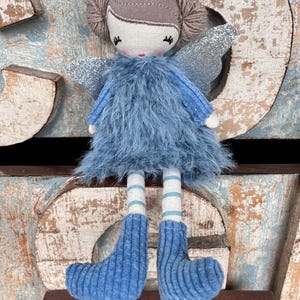 Peut inclure: Poup&eacute;e f&eacute;e faite &agrave; la main, v&ecirc;tue d'une robe et de bottes bleu clair duveteuses. La poup&eacute;e a des ailes argent&eacute;es scintillantes, une t&ecirc;te marron clair et des chaussettes ray&eacute;es. La poup&eacute;e est pos&eacute;e sur une surface marron avec un embl&egrave;me dor&eacute;.