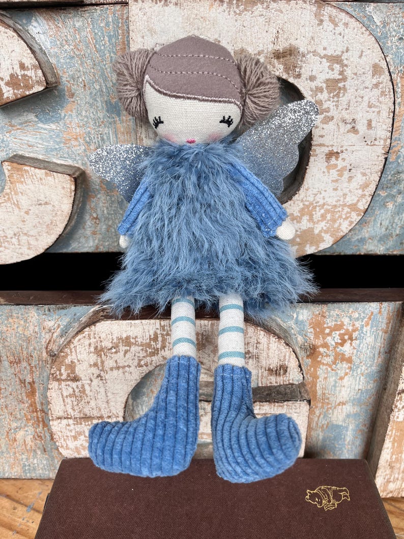 Peut inclure: Poup&eacute;e f&eacute;e faite &agrave; la main, v&ecirc;tue d'une robe et de bottes bleu clair duveteuses. La poup&eacute;e a des ailes argent&eacute;es scintillantes, une t&ecirc;te marron clair et des chaussettes ray&eacute;es. La poup&eacute;e est pos&eacute;e sur une surface marron avec un embl&egrave;me dor&eacute;.