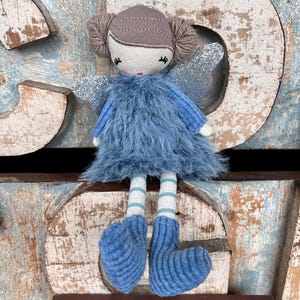 Peut inclure: Une poup&eacute;e f&eacute;e faite &agrave; la main avec une robe en fourrure bleue, des ailes en paillettes argent&eacute;es et des jambes ray&eacute;es. La poup&eacute;e a une t&ecirc;te beige avec des cheveux en fil marron et des bottes bleues tricot&eacute;es.