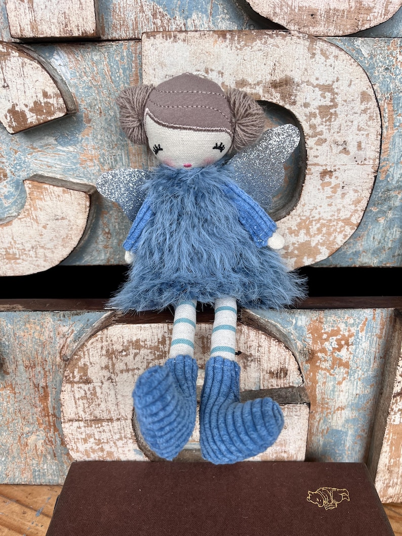 Peut inclure: Une poup&eacute;e f&eacute;e faite &agrave; la main avec une robe en fourrure bleue, des ailes en paillettes argent&eacute;es et des jambes ray&eacute;es. La poup&eacute;e a une t&ecirc;te beige avec des cheveux en fil marron et des bottes bleues tricot&eacute;es.
