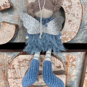 Peut inclure: Une poup&eacute;e f&eacute;e faite &agrave; la main avec des ailes en paillettes argent&eacute;es, une jupe en fourrure bleue et des bottes en tricot bleu. La poup&eacute;e a un visage beige, des couettes en fil marron et des jambes ray&eacute;es. Le fond pr&eacute;sente des lettres en bois vieilli.