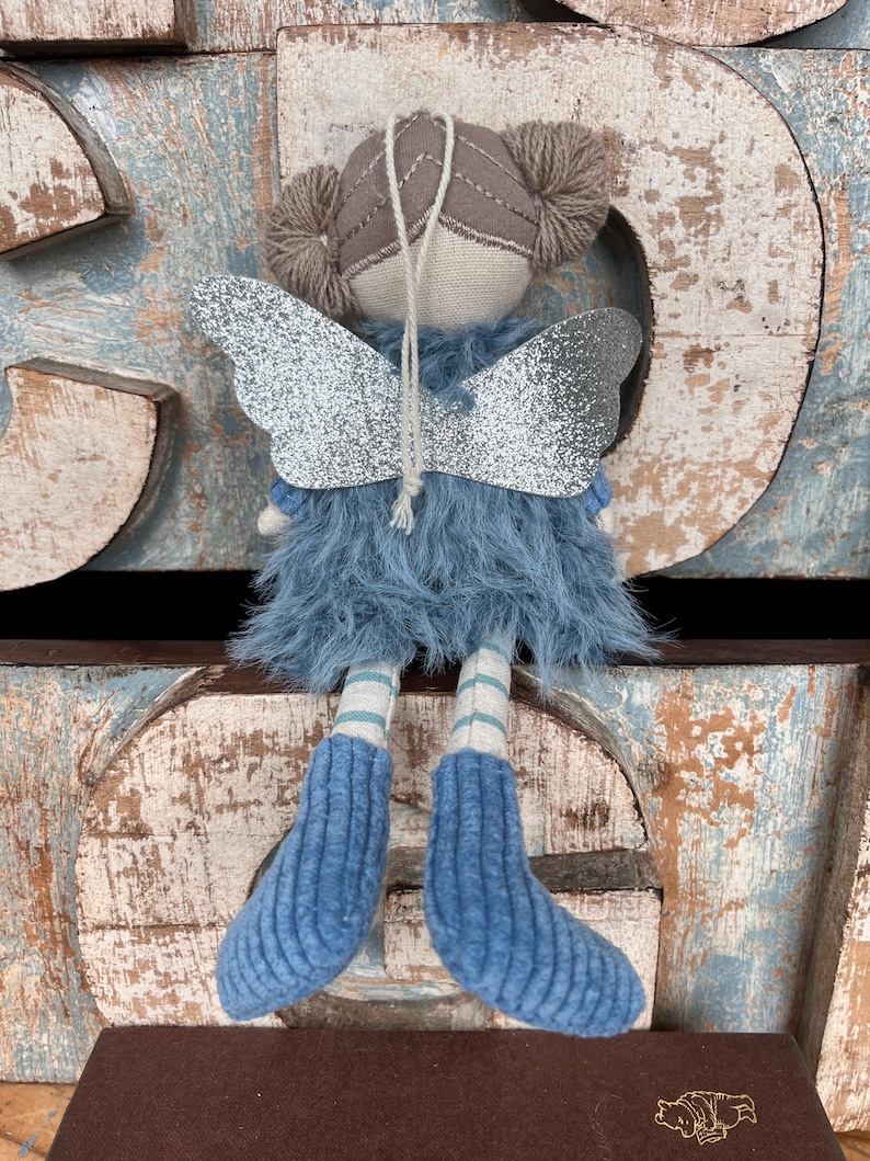 Peut inclure: Une poup&eacute;e f&eacute;e faite &agrave; la main avec des ailes en paillettes argent&eacute;es, une jupe en fourrure bleue et des bottes en tricot bleu. La poup&eacute;e a un visage beige, des couettes en fil marron et des jambes ray&eacute;es. Le fond pr&eacute;sente des lettres en bois vieilli.