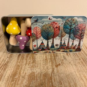 May include: A rectangular metal tin containing several small wooden mushroom figures in yellow, purple, and red with white spots. The lid of the tin features a colourful illustration of stylised trees and small dwellings under a cloudy sky.