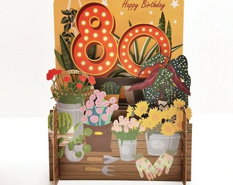 Puede incluir: Una tarjeta de cumpleaños 3D con una ilustración colorida de una floristería. La tarjeta presenta un gran letrero iluminado con el número 80 y las palabras "Feliz cumpleaños".