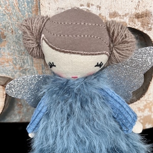 Peut inclure: Une poup&eacute;e f&eacute;e faite &agrave; la main avec une robe en plumes bleu clair et des ailes en paillettes argent&eacute;es. La poup&eacute;e a un visage beige avec les yeux ferm&eacute;s et une bouche rose. Les cheveux sont bruns et coiff&eacute;s en deux chignons.