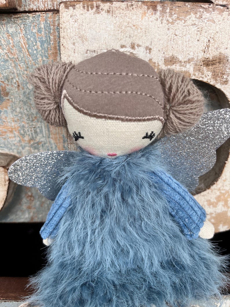 Peut inclure: Une poup&eacute;e f&eacute;e faite &agrave; la main avec une robe en plumes bleu clair et des ailes en paillettes argent&eacute;es. La poup&eacute;e a un visage beige avec les yeux ferm&eacute;s et une bouche rose. Les cheveux sont bruns et coiff&eacute;s en deux chignons.