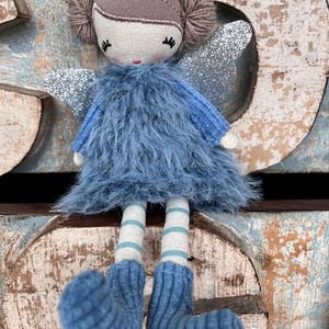 Peut inclure: Une poup&eacute;e f&eacute;e faite &agrave; la main avec une robe en fourrure bleue, des ailes en paillettes argent&eacute;es et des jambes ray&eacute;es. La poup&eacute;e a une t&ecirc;te brune avec des couettes en fil et un visage peint. Les pieds bleus tricot&eacute;s de la poup&eacute;e sont visibles.