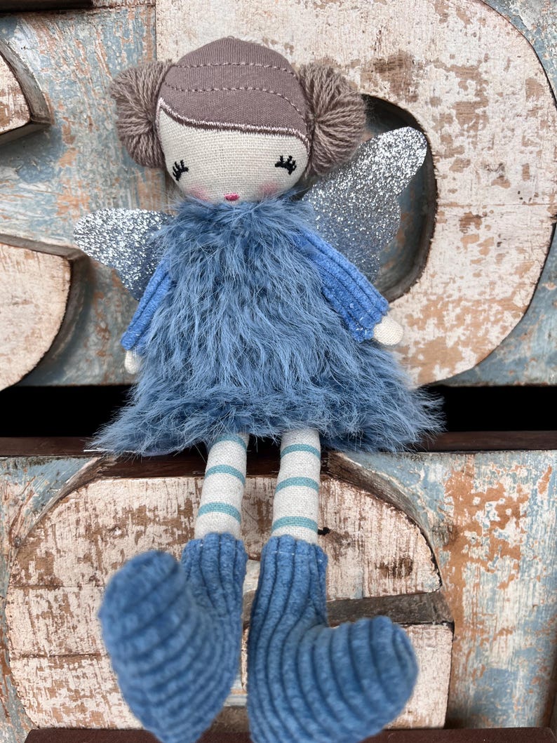 Peut inclure: Une poup&eacute;e f&eacute;e faite &agrave; la main avec une robe en fourrure bleue, des ailes en paillettes argent&eacute;es et des jambes ray&eacute;es. La poup&eacute;e a une t&ecirc;te brune avec des couettes en fil et un visage peint. Les pieds bleus tricot&eacute;s de la poup&eacute;e sont visibles.