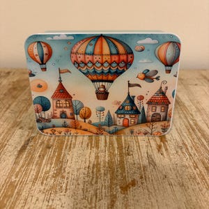 May include: A rectangular metal tin featuring a colourful illustration of three hot air balloons floating above a whimsical village with small houses, trees, and a bird against a light blue sky with soft clouds and a warm, orange-toned horizon.