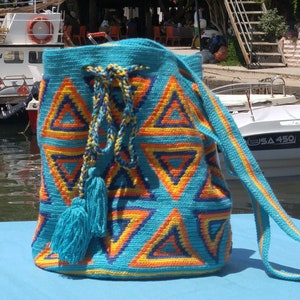 May include: A turquoise crocheted bag with a geometric pattern of orange, yellow, and blue triangles. The bag has a long turquoise strap and a turquoise tassel.