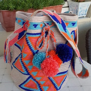 May include: A crocheted tote bag with a geometric pattern in shades of blue, orange, and pink. The bag has a long strap and a button closure. It is decorated with pom-poms in blue, orange, and pink.