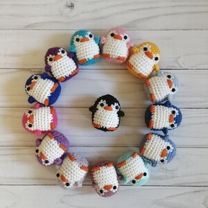 May include: A circle of crocheted penguin ornaments in various colors, including blue, purple, pink, yellow, and white. The penguins are arranged in a circle around a black and white penguin in the center.