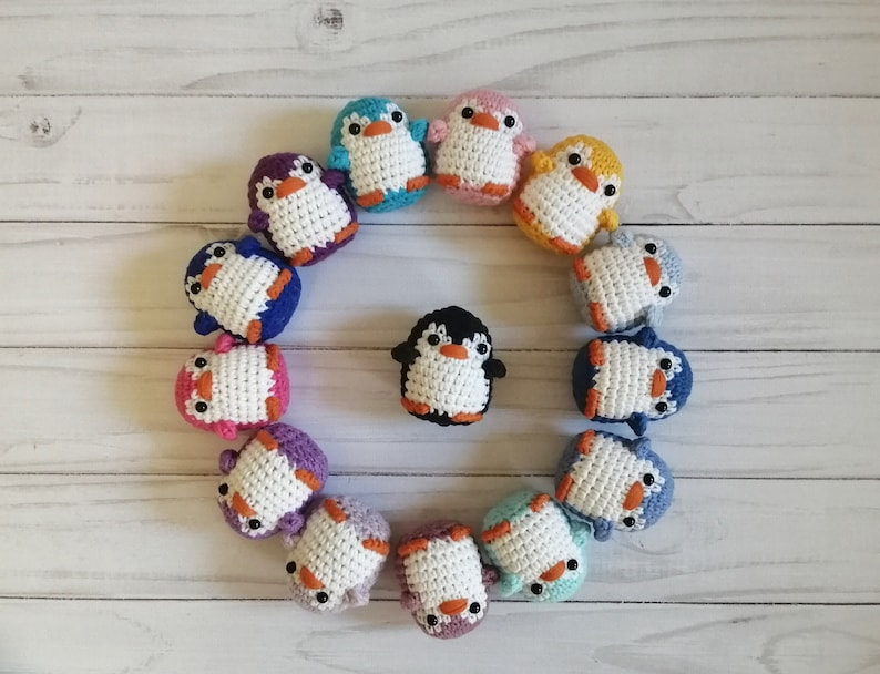 May include: A circle of crocheted penguin ornaments in various colors, including blue, purple, pink, yellow, and white. The penguins are arranged in a circle around a black and white penguin in the center.