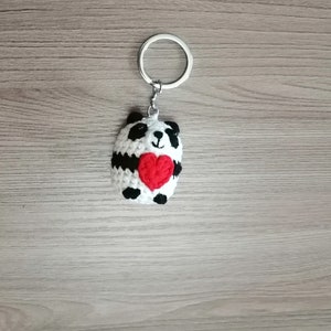 May include: A white and black crocheted panda bear keychain holding a red heart. The keychain is attached to a silver metal ring.