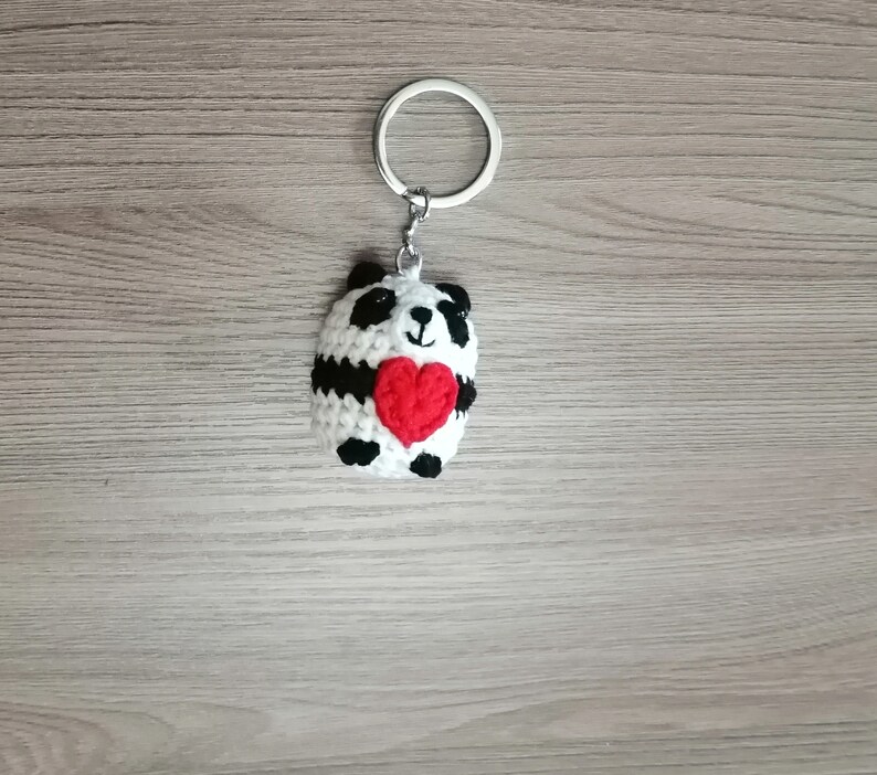 May include: A white and black crocheted panda bear keychain holding a red heart. The keychain is attached to a silver metal ring.