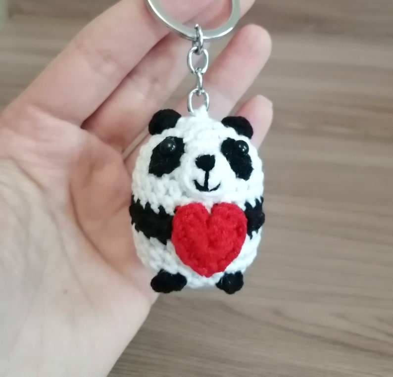 May include: A white crocheted panda bear keychain holding a red heart. The panda has black eyes and a black nose.