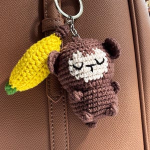 May include: Crocheted monkey keychain with a yellow banana. The monkey is brown with a white face and closed eyes. The banana has green accents. The keychain is attached to a silver ring.