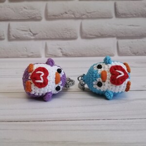 May include: Two crocheted penguin keychains with white bodies, orange beaks, black eyes, and red hearts on their chests. One penguin is purple and the other is blue.