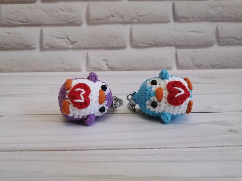 May include: Two crocheted penguin keychains with white bodies, orange beaks, black eyes, and red hearts on their chests. One penguin is purple and the other is blue.