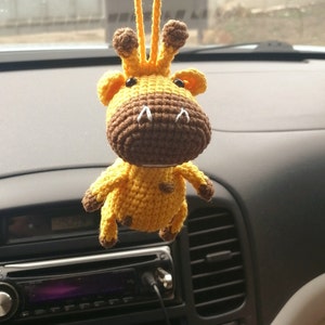 May include: A crocheted giraffe toy with brown and yellow yarn. The giraffe has a long neck and a small head with brown eyes and a white nose. The toy is hanging from a rearview mirror in a car.