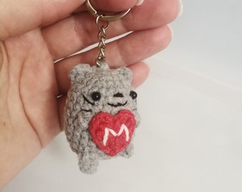 May include: Gray crocheted cat keychain holding a red heart with a white letter M on it.