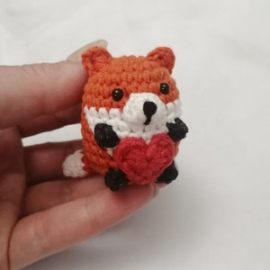 Puede incluir: Un pequeño zorro de crochet sosteniendo un corazón rojo. El zorro es naranja con detalles blancos y ojos negros.