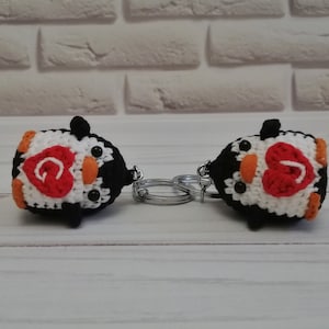 May include: Two crocheted penguin keychains with black and white bodies, orange beaks, and black eyes. Each penguin has a red heart on its chest with a letter "G" inside.