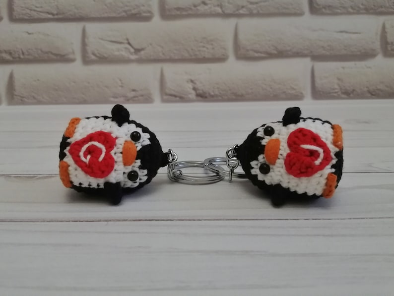 May include: Two crocheted penguin keychains with black and white bodies, orange beaks, and black eyes. Each penguin has a red heart on its chest with a letter "G" inside.