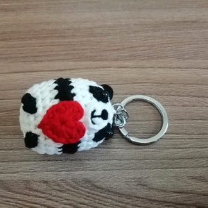 May include: A crocheted panda keychain holding a red heart. The keychain is attached to a silver metal ring.