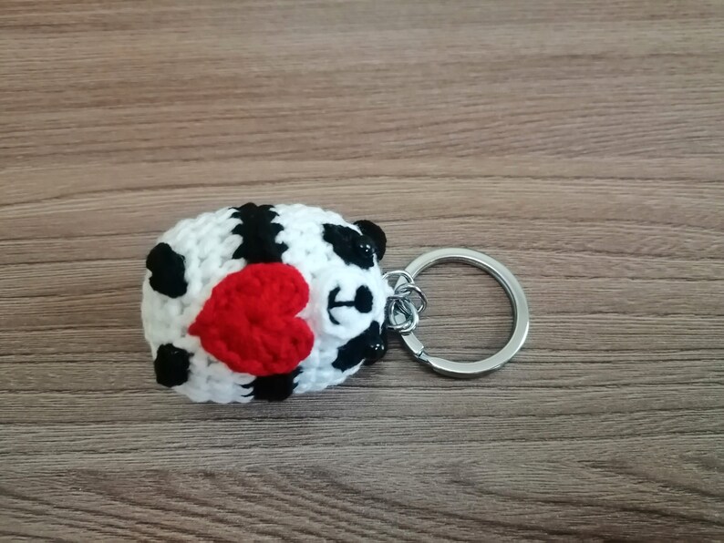 May include: A crocheted panda keychain holding a red heart. The keychain is attached to a silver metal ring.