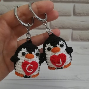 May include: Two crocheted penguin keychains with red hearts. The hearts have the letters "G" and "J" on them.