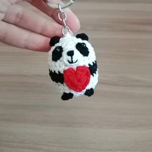 May include: A white and black crocheted panda bear keychain holding a red heart. The keychain is attached to a silver key ring.