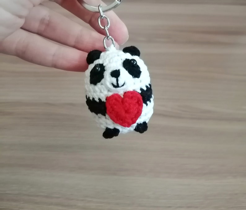 May include: A white and black crocheted panda bear keychain holding a red heart. The keychain is attached to a silver key ring.