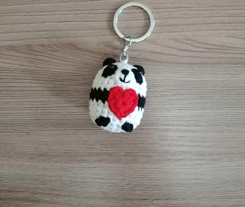 May include: A white crocheted panda bear keychain holding a red heart. The keychain is attached to a silver key ring.
